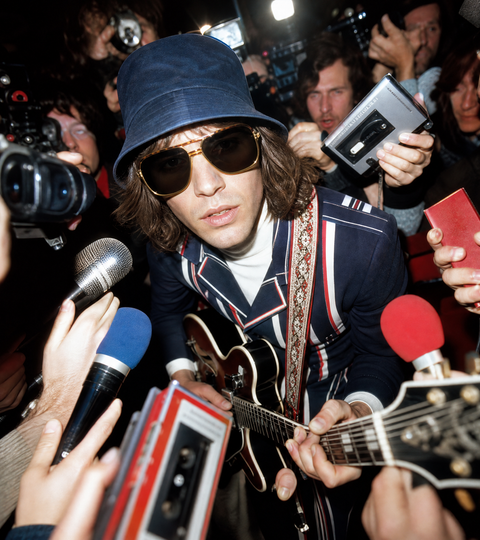 Man wearing a bucket hat and sunglasses playing the guitar for the press in mod clothing